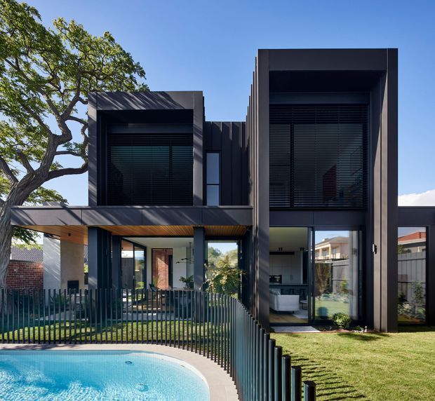 Exterior view of a contemporary multi-level residence with a bold dark facade, projecting rectangular forms, extensive glazing, and deep window reveals. A covered outdoor area and balcony sit beside a fenced swimming pool, with mature landscaping and neighboring homes visible around the property.