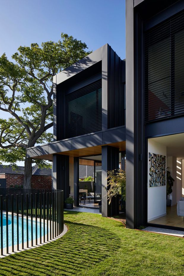 A contemporary two-storey residential house viewed from the landscaped backyard. The dark, rectilinear facade features projecting upper-level volumes, large windows with horizontal blinds, deep overhangs, and vertical metal detailing. A partially visible swimming pool is enclosed by a slim black vertical-bar fence, with lawn, mature trees, and a covered outdoor seating area around the house. Interior artwork and furnishings are visible through the open ground-floor glazing.