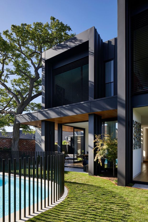 Exterior view of a contemporary multi-level residence with dark grey rendered walls, strong geometric forms, large shaded windows, timber soffit lining and extensive glazing. A swimming pool with black vertical-bar fencing occupies the foreground, with landscaped lawn and mature trees surrounding the house.