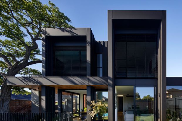 Contemporary multi-level residential facade with strong geometric volumes, dark framed glazing, deep recessed windows, and a sheltered entry or outdoor living area. The exterior is finished in dark grey panels with contrasting lighter grey projecting frames, set beside mature landscaping and neighbouring homes.