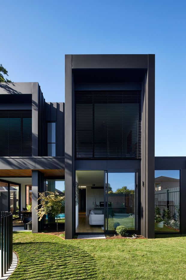 Exterior architectural photograph of a contemporary multi-level residential home. The facade features tall dark-framed glazing, strong rectangular projecting frames, horizontal exterior blinds, dark cladding, and a timber-lined soffit over a sheltered outdoor area. A lawn and landscaped garden sit in the foreground, with living spaces visible through the glass.