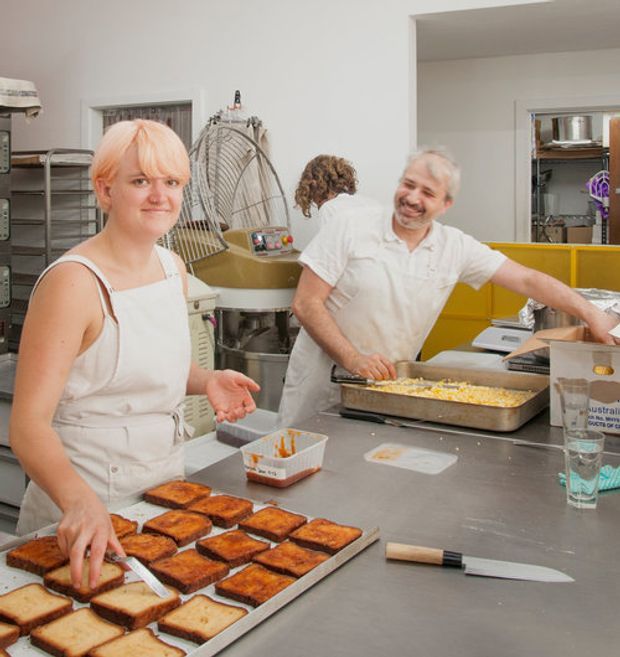 A bright commercial food preparation space, likely a bakery or professional kitchen, with two people working at a large stainless-steel workbench. Trays of baked goods, mixing equipment, shelving, and food-preparation tools are visible. The room has white walls with a bold yellow tiled or painted backsplash and utilitarian stainless-steel surfaces.