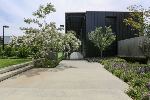 A contemporary dark-clad building viewed from a landscaped pedestrian approach. The facade features vertical black cladding, large dark-framed glazing, and a covered recessed entry walkway. Concrete paving, raised steps, garden beds, ornamental grasses, small trees, and a weathered metal water feature define the foreground.