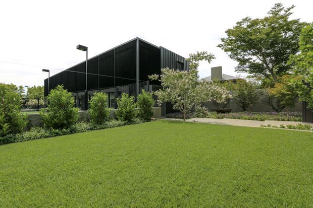 A contemporary black-clad building viewed from a landscaped lawn. The angular structure features dark vertical metal cladding, large glazed openings, simple rectangular volumes, and minimalist black exterior lighting. Mature trees, shrubs, paved paths, and a manicured grass foreground surround the building.