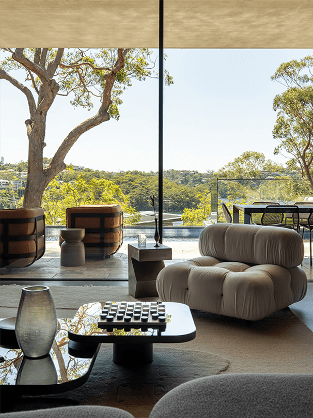 A sophisticated contemporary living room with sculptural furniture arranged beside floor-to-ceiling glazing. The space features a plush cream modular armchair, dark reflective coffee table, geometric side table, textured rug, and warm neutral finishes. A covered outdoor terrace, pool edge, mature trees, and a green hillside are visible beyond the glazing.