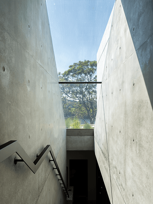 A narrow, dramatic stairwell or passage between tall exposed concrete walls, with a zigzag metal handrail on the left and a dark stair descending toward an opening. A slender glazed or open slot at the far end frames a mature tree and bright blue sky, while a thin black structural bridge spans the upper opening. The composition emphasizes raw concrete, strong vertical geometry, natural light, and a sculptural architectural design.