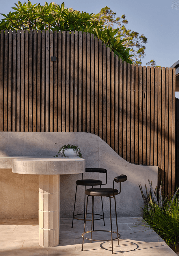 A warm, sunlit outdoor entertaining area with a curved rendered concrete or plaster bar counter and integrated curved bench, paired with two black metal bar stools. The space is enclosed by a tall vertical timber slat privacy fence, with paving underfoot, ornamental grasses, and lush tropical planting above the fence. A partial roof or structure is visible at the right edge.