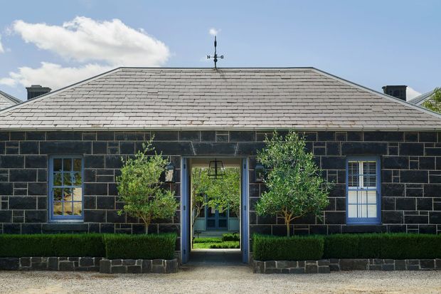 Front elevation of a single-storey symmetrical house with dark masonry block walls, a low-pitched hipped slate roof, blue-trimmed windows, and a central glazed entry. Formal clipped hedges and small ornamental trees frame the entrance, with a gravel forecourt in front.
