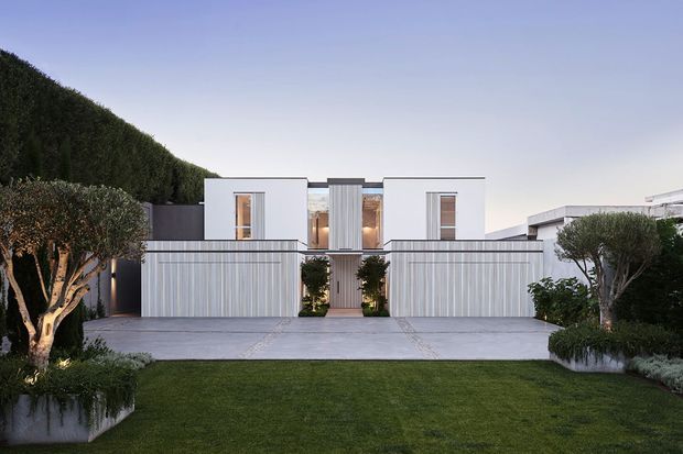 A symmetrical contemporary residential facade viewed from the street, featuring crisp white rendered cubic volumes, tall narrow windows, a central recessed timber-toned entry, and matching vertical slatted garage or boundary doors. A broad concrete driveway, manicured lawn, clipped hedges, and sculpted trees create a highly ordered landscaped frontage.