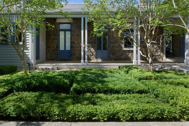 A single-storey building facade viewed from a landscaped garden, featuring a covered verandah with slender white posts, dark blue-grey doors and window frames, pale horizontal weatherboard cladding, and prominent rough stone masonry wall sections. Dense clipped green hedging fills the foreground.