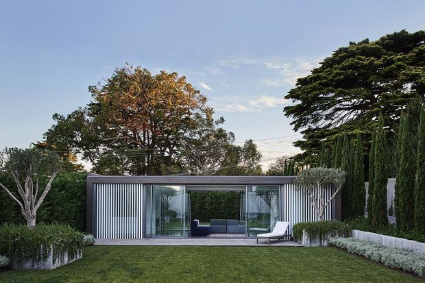 Contemporary single-storey garden pavilion with a flat roof, extensive floor-to-ceiling glazing, and vertical metal screening. The compact structure opens onto a manicured lawn and is framed by sculpted trees, tall hedging, and raised planting beds. A minimal lounge arrangement is visible inside and on the terrace.