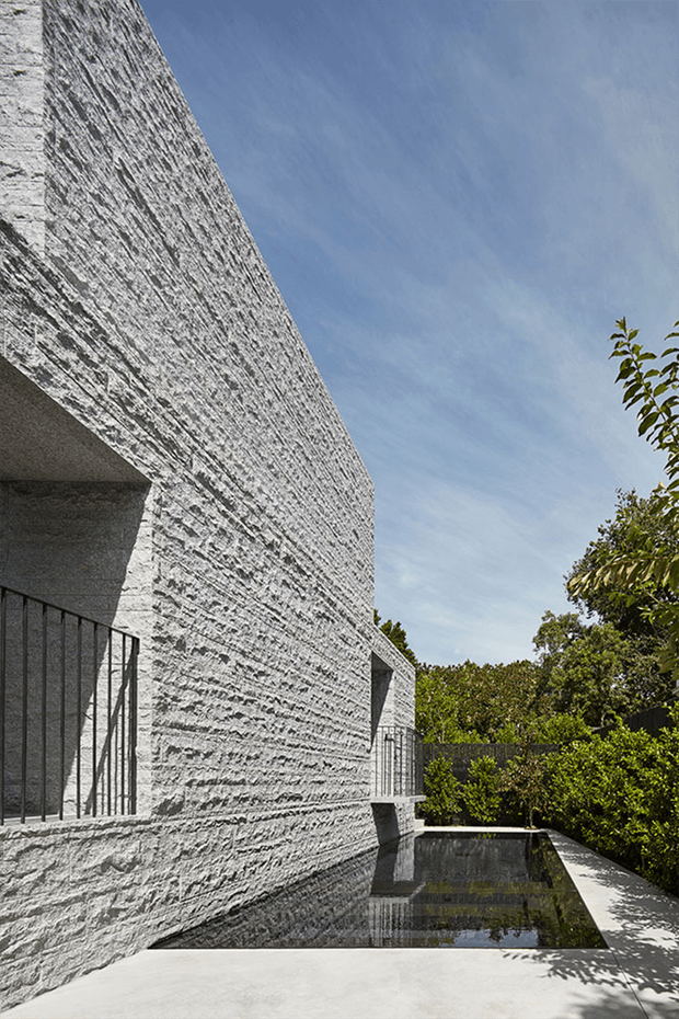 A dramatic exterior architectural view of a sculptural, monolithic building clad in heavily textured light-grey stone or concrete masonry. Deep recessed openings and black metal balcony railings create strong shadow lines, while a narrow reflecting pool runs alongside the building. Dense garden planting and a bright blue sky frame the contemporary composition.