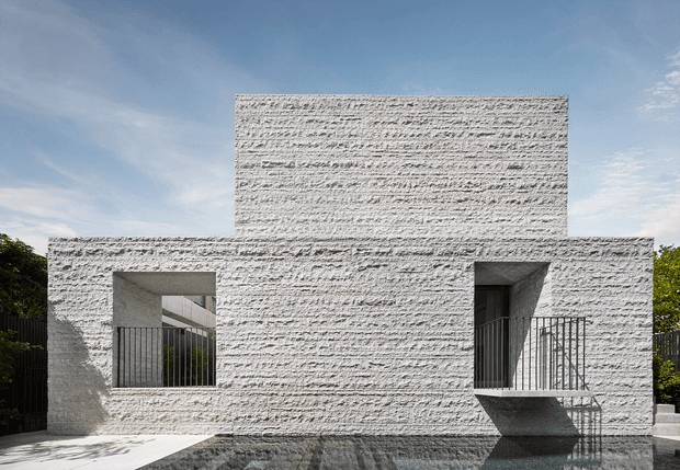 A minimalist modern residential facade composed of strong rectilinear volumes clad in heavily textured white masonry or render. Deep recessed openings, dark metal balustrades, and sharp geometric shadows create a sculptural, high-contrast exterior against a clear blue sky.