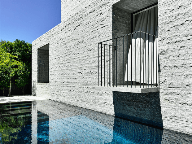 Exterior view of a minimalist contemporary residence with a heavily textured light-grey masonry facade, deep-set openings, and a projecting upper-level balcony enclosed by slender black metal railings. A reflective swimming pool runs alongside the building, mirroring the facade and blue sky, with mature greenery visible at the edge.