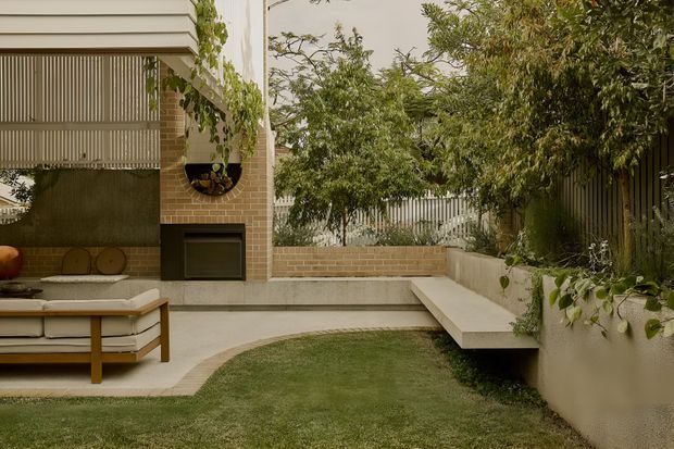A landscaped residential courtyard entertaining area with a built-in outdoor fireplace set into a warm brick wall, a concrete-paved terrace, low integrated bench seating, and a partially visible outdoor sofa. Vertical screening, lush planting, and mature trees create a private garden setting. The composition has a refined contemporary character with warm neutral materials.