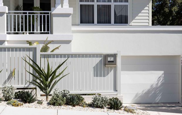 Bright daytime view of a white residential facade with a garage door, upper-level window, balcony railing, entry gate, mailbox, and drought-tolerant landscaping with succulents and gravel.