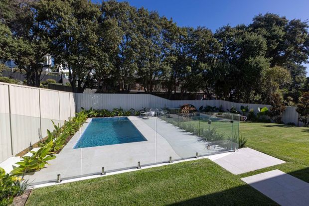 A bright landscaped backyard with a rectangular in-ground swimming pool set into a light concrete-paved terrace. Frameless glass pool fencing surrounds the pool, with a manicured lawn, stepping-stone path, planted garden beds, and a white boundary fence beneath mature trees.