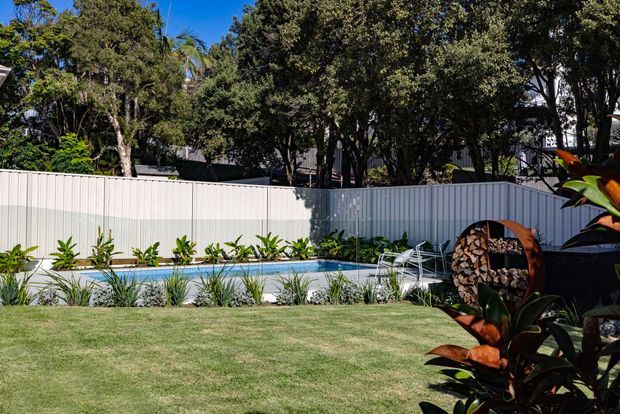 A landscaped backyard with a narrow rectangular swimming pool bordered by a light-coloured metal privacy fence, lawn, tropical planting, and mature trees. A sculptural outdoor firewood feature is visible in the foreground.