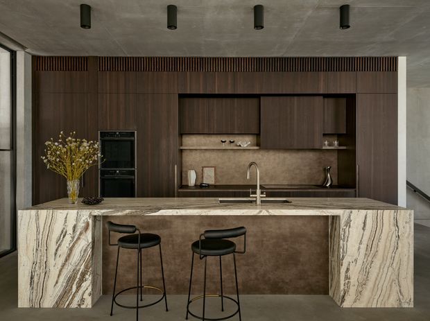 A sophisticated contemporary kitchen with a large sculptural stone island and integrated breakfast bar. The kitchen features dark timber cabinetry, a mottled stone or plaster-look splashback, black appliances, a stainless steel tap and sink, black bar stools, and a raw concrete ceiling with recessed cylindrical lights. Full-height glazing is visible at the left and right edges.