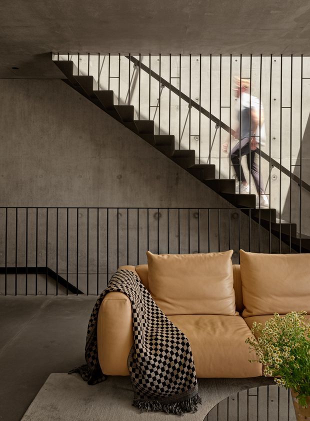 A moody contemporary interior featuring a concrete stairway with slim black metal balustrades, viewed behind a tan leather sofa. A black-and-cream geometric throw, soft rug, round coffee table, and greenery add warmth to the raw concrete setting. A blurred person is visible on the stairs.