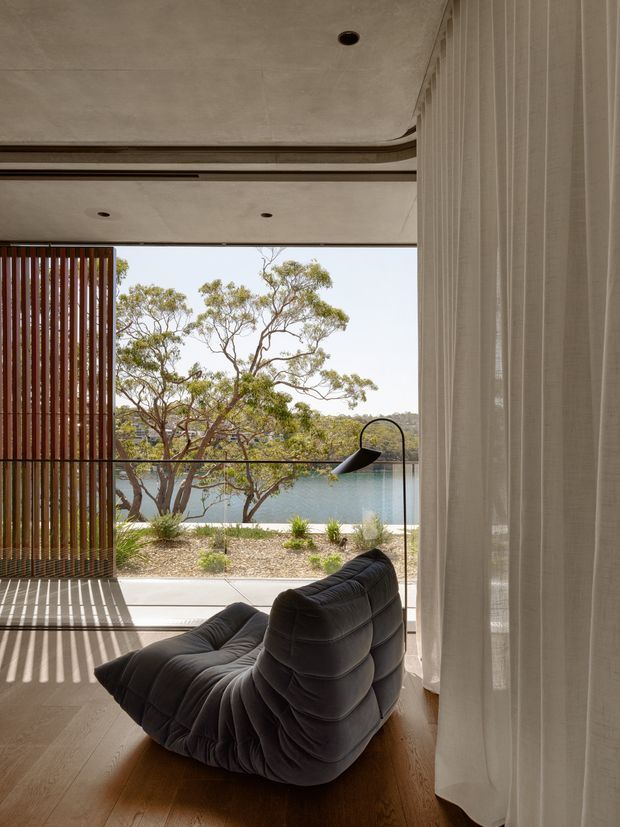 A refined living room opening onto a landscaped waterfront setting. The room features a low, sculptural dark modular sofa, pale timber flooring, sheer floor-to-ceiling curtains, a black arched floor lamp, and a concrete ceiling with recessed downlights. Vertical timber screening creates striped sunlight and shade near the open glazing, with water and trees visible beyond.