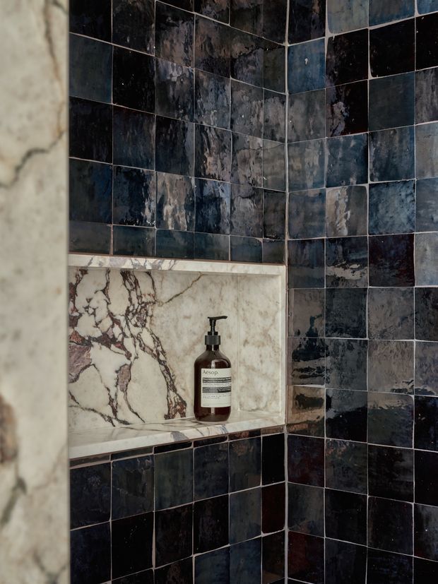 Close-up of a tiled bathroom shower or bathing area. Dark blue-black, heavily variegated square wall tiles cover the corner, with a recessed marble niche containing a pump bottle. The niche is framed with pale veined stone, creating a dramatic, moody material contrast.