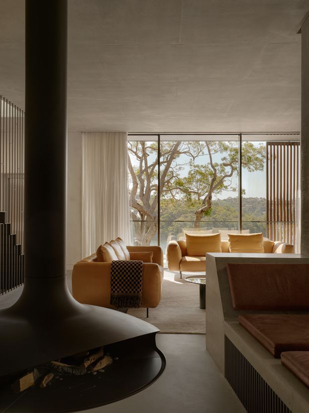 A warm, sophisticated living room viewed from beside a large sculptural black freestanding fireplace. The room has a smooth pale concrete or plaster ceiling and floor, expansive floor-to-ceiling glazing overlooking mature trees, sheer curtains, caramel and tan upholstered lounge seating, and a built-in bench or joinery element in the foreground. Vertical dark slatted detailing frames the fireplace and foreground cabinetry, creating a refined contemporary interior with strong indoor-outdoor connection.