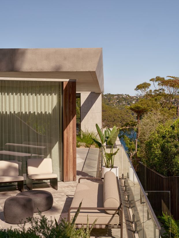 A contemporary outdoor terrace beside a minimalist concrete and timber-clad building. The terrace features a glass balustrade, pale tiled or concrete paving, a cushioned outdoor sofa, sculptural lounge chairs, a low ottoman, and potted tropical plants. Floor-to-ceiling glazing opens to the interior, while a leafy suburban hillside and glimpse of blue water are visible beyond.