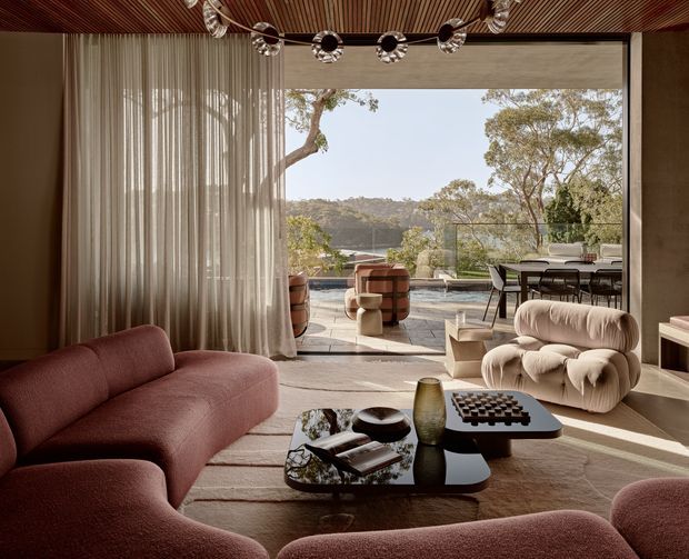 Warm, refined living room with a curved dusty-rose sofa, sculptural cream lounge chair, glass coffee table, layered rug, sheer curtains, and a timber slatted ceiling. Full-height glazing opens to a furnished terrace with views toward a wooded hillside and water. A sculptural multi-light chandelier and dark timber, stone, and neutral finishes create a luxurious contemporary interior.
