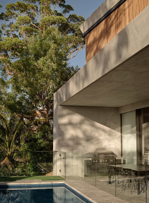 Contemporary residential exterior with a cantilevered concrete terrace or overhang beside a swimming pool. The facade combines smooth exposed concrete with a vertical timber-screen feature and large glazed openings. A covered outdoor dining and barbecue area is visible behind frameless glass, surrounded by dense established greenery and mature trees. Warm late-afternoon light creates strong organic shadows across the concrete surfaces.