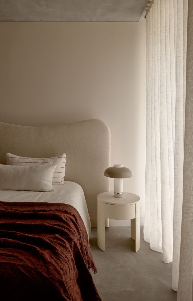 A softly lit, minimalist bedroom corner featuring a curved upholstered bedhead, white linen bedding, a deep rust-brown throw, a small round bedside table with a sculptural mushroom lamp, and full-height sheer curtains. The room has warm neutral walls and a smooth concrete-like floor and ceiling, creating a calm, refined atmosphere.