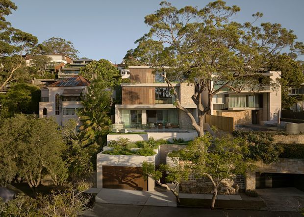 Exterior view of a contemporary multi-level residential development set among mature trees on a sloping suburban site. The homes feature flat rooflines, rectilinear forms, large glazed openings, timber-clad upper sections, light masonry or rendered walls, landscaped terraces, and integrated garages. The image primarily presents the street-facing facades and surrounding garden setting.