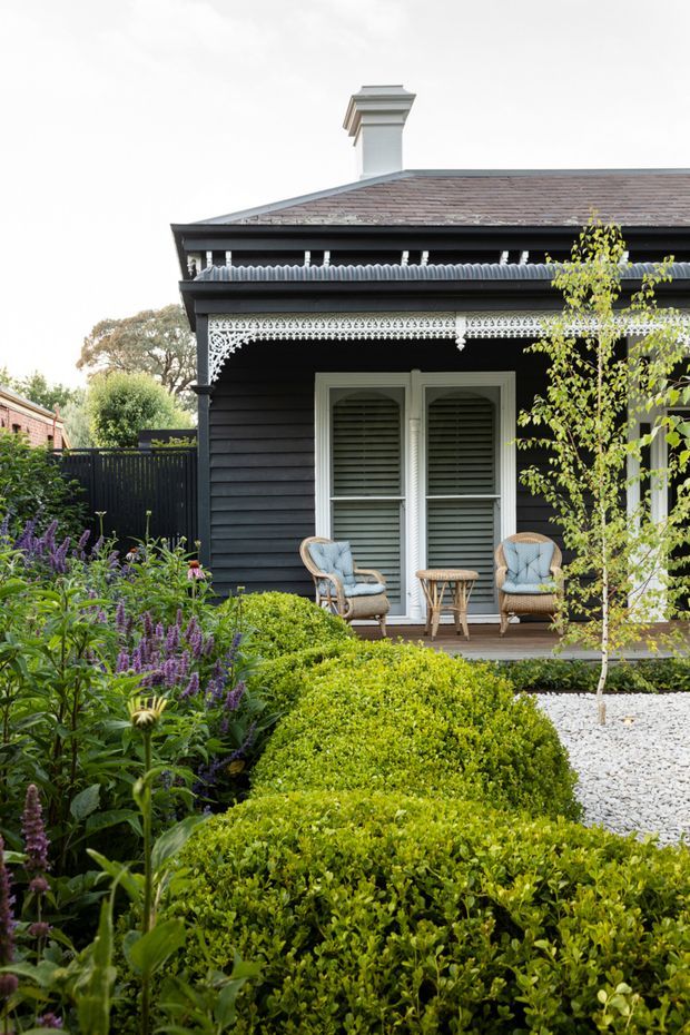 Exterior view of a heritage-style dark weatherboard house with ornate decorative veranda trim, a hipped tiled roof and chimney. White French doors with louvred shutters open onto a small timber porch furnished with two wicker armchairs and a small round table. The foreground is a carefully designed garden with clipped hedges, flowering plants, a pale gravel path and a young tree.