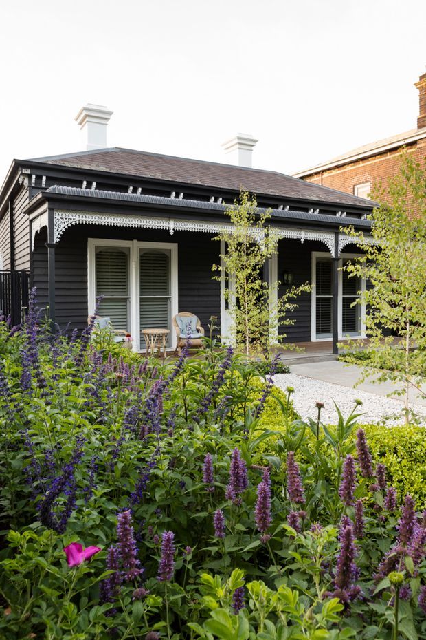 A single-storey heritage-style cottage with dark horizontal weatherboard cladding, a low hipped roof, decorative white verandah fretwork, white chimneys and shuttered windows. It is viewed from a lush, densely planted garden with purple flowering plants, gravel paths and clipped greenery. A larger brick building is visible behind the cottage.