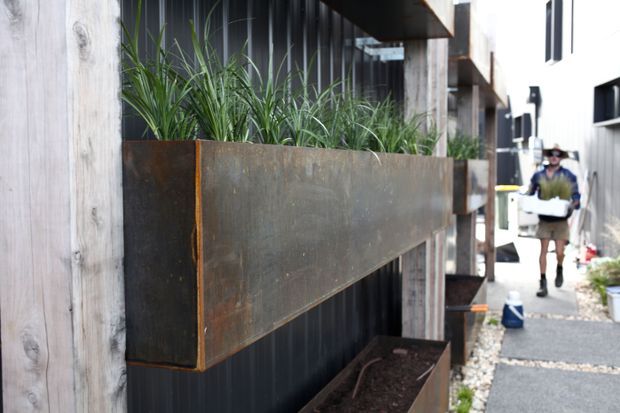 Close-up exterior view of a contemporary building facade featuring large rusted steel planter boxes mounted against black corrugated cladding and framed by vertical timber posts. A narrow gravel-paved walkway and neighbouring building are visible to the right.