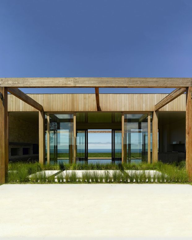 Front-facing exterior view of a contemporary pavilion-like residence with exposed timber posts and beams, extensive floor-to-ceiling glazing, and open-plan interiors visible beyond. A landscaped planted strip separates the building from a broad pale concrete forecourt, while a blue ocean horizon is visible through the central glazing.