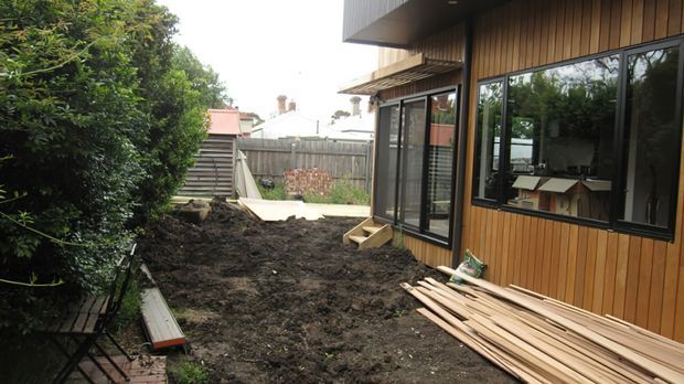 Side and rear exterior view of a contemporary residential extension under construction. The building has vertical timber cladding, dark-framed glazing and a projecting upper roof or soffit, with exposed soil and stacks of timber boards in the narrow side yard. A fenced suburban backyard and neighboring homes are visible beyond.