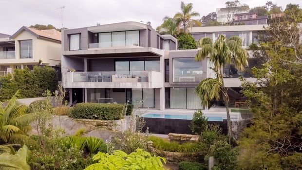 Exterior view of a multi-storey contemporary residential home on a landscaped, sloping site. The building has a dark rendered facade, extensive floor-to-ceiling glazing, projecting balcony slabs, and a rectangular swimming pool in front, with neighbouring homes and tropical planting visible around it.