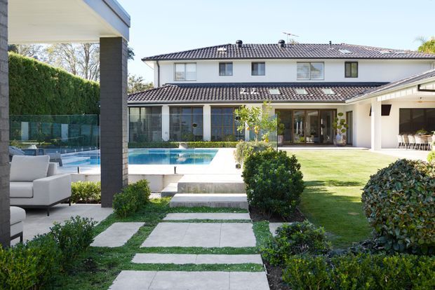 A spacious landscaped courtyard of a large two-storey suburban home, viewed from a covered outdoor lounge. The scene features a rectangular swimming pool, manicured lawn and hedges, concrete stepping-stone paths, dark tiled pitched roofs, pale rendered walls, extensive glazing, and a covered outdoor dining area.