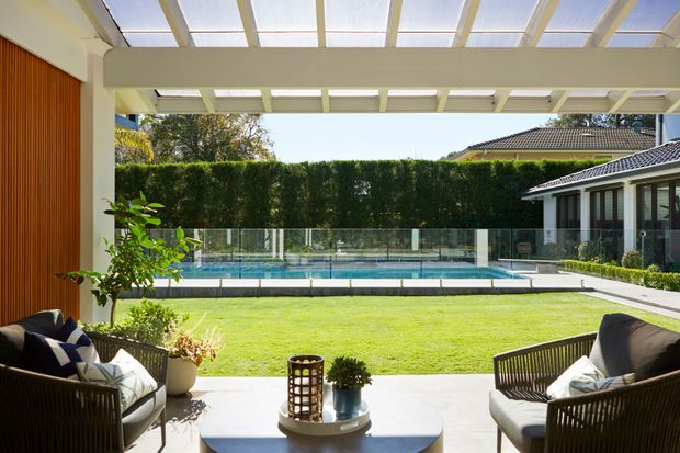 A bright covered outdoor living area opens onto a manicured lawn and long swimming pool. The space features white structural beams and translucent roof panels, timber slat wall cladding, glass pool fencing, and woven lounge chairs with cushions. A neighboring or adjoining pavilion structure is visible to the right.