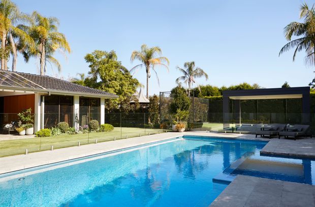 A bright landscaped backyard with a large rectangular swimming pool, paved surrounds, and a covered outdoor lounge pavilion. The pool area is enclosed by frameless glass fencing, with a single-storey tiled-roof house and lush palm planting in the background.