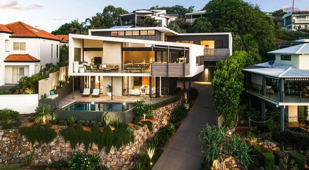 Exterior view of a contemporary multi-level residence on a sloping site, featuring strong horizontal volumes, white rendered walls, dark timber-clad sections, extensive glazing, balconies, and a swimming pool terrace. The landscaped frontage includes stone retaining walls and dense subtropical planting, with neighbouring homes visible on either side.