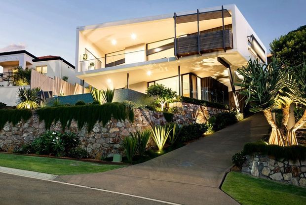 A modern multi-level residential house photographed from the street at dusk. The white rendered facade features broad open balconies, extensive glazing, dark horizontal privacy screens, black-framed windows and doors, and warm architectural lighting. A sloping concrete driveway leads to the entry, bordered by layered stone retaining walls and drought-tolerant landscaped planting.