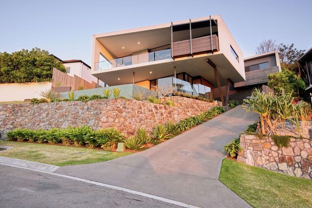 Contemporary multi-level residential house on a sloped suburban site, featuring a projecting flat-roofed upper volume, broad glazed balconies, timber privacy screens, pale rendered walls, and extensive natural-stone retaining walls with landscaped gardens. A concrete driveway rises toward the house from the street.