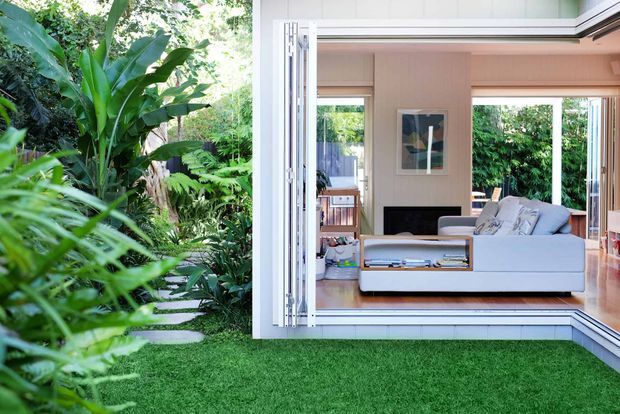 A bright contemporary living room opens directly onto a lush tropical garden through fully retractable glazed folding doors. The room features a pale upholstered sectional sofa, built-in timber-toned shelving, a low fireplace or dark media niche, pale walls, and warm timber flooring. Dense greenery and a manicured lawn frame the indoor-outdoor connection.