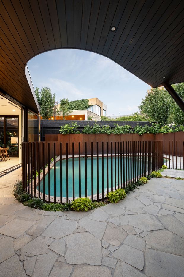 A landscaped residential courtyard viewed from beneath a curved timber-lined canopy. The scene features a small turquoise swimming pool enclosed by a dark vertical metal balustrade, irregular natural stone paving, rust-toned boundary fencing, planted garden beds, and glimpses of a modern home and dining area beyond.