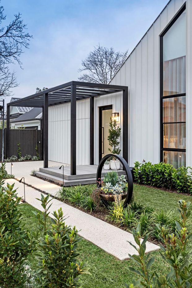 A landscaped residential exterior with a contemporary white-panelled facade, tall black-framed glazing, and a dark steel pergola over a raised entry deck. A pale concrete path leads through manicured lawn and layered planting toward the entrance, with a sculptural circular garden planter as a feature.