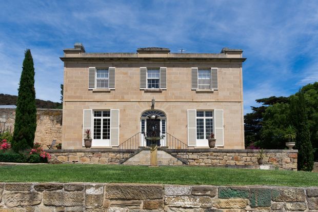 Front exterior view of a symmetrical two-storey stone residence with a flat parapet roof, tall shuttered windows, classical proportions, central arched entry, and formal landscaped garden terraces. The facade and retaining walls are constructed in warm beige natural stone, with dark green cypress and clipped planting framing the building.