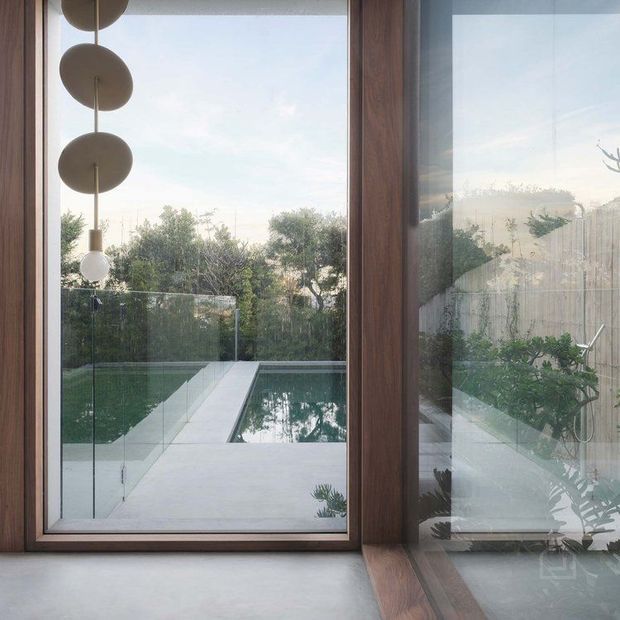 Interior view through expansive floor-to-ceiling glazing toward a compact rectangular swimming pool and paved terrace. A warm timber-framed glazed door or window dominates the foreground, with a sculptural pendant light at left and reflections revealing the room. The outdoor area includes a frameless glass balustrade, landscaped planting, and a timber privacy screen.