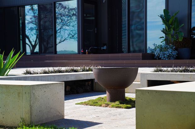 A dark, contemporary building entrance opens onto a minimalist concrete courtyard. Broad concrete benches or plinths frame a paved area with a large rust-toned bowl planter or sculptural feature, low planting, and deep timber-toned steps. Full-height glazing reflects surrounding trees and landscape, while architectural planting adds greenery.